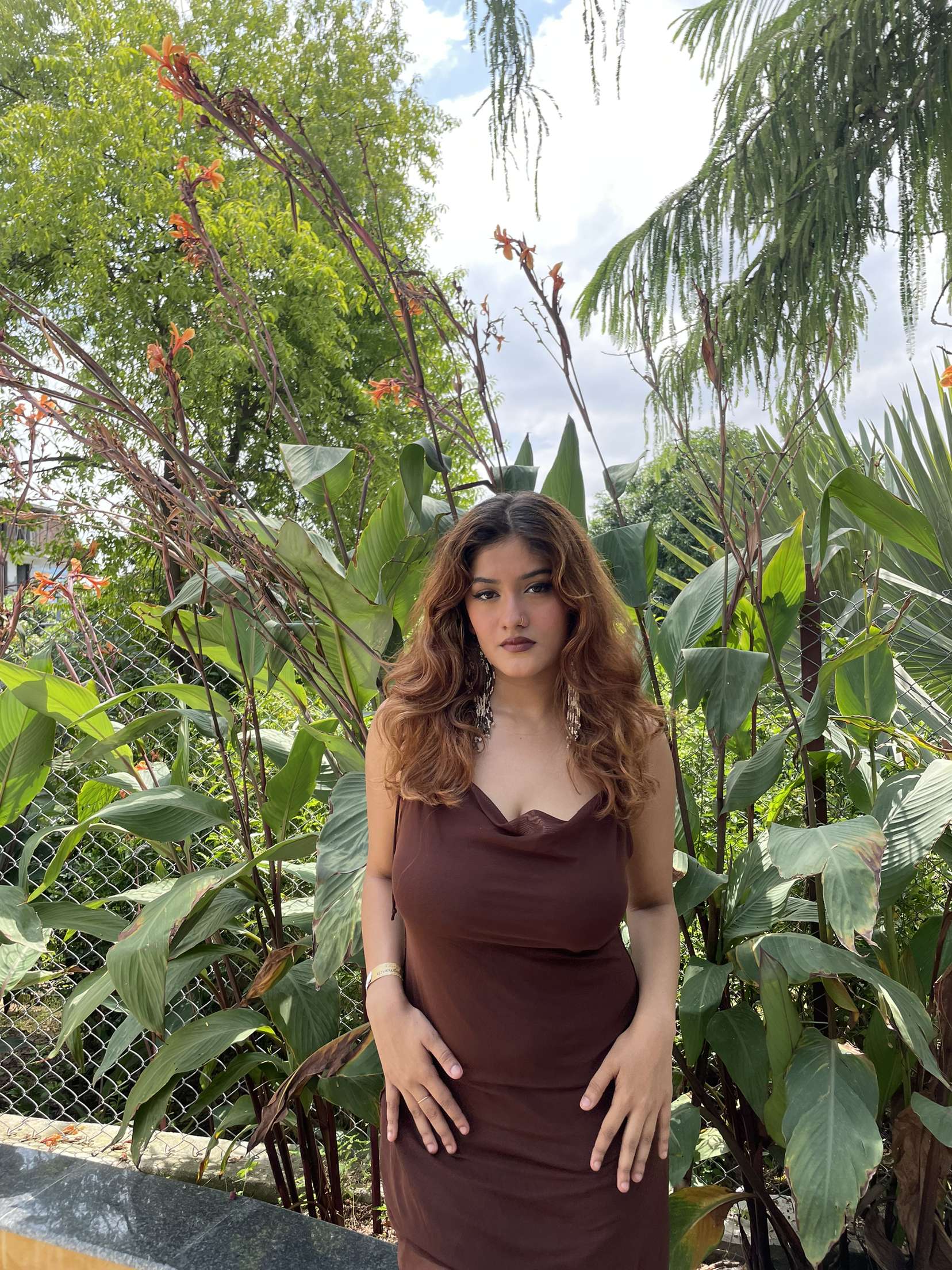 Woman in a brown dress posing beside flowering plants and tall foliage in the garden at Rangmanch Farms, an outdoor event venue in Gurugram near Delhi