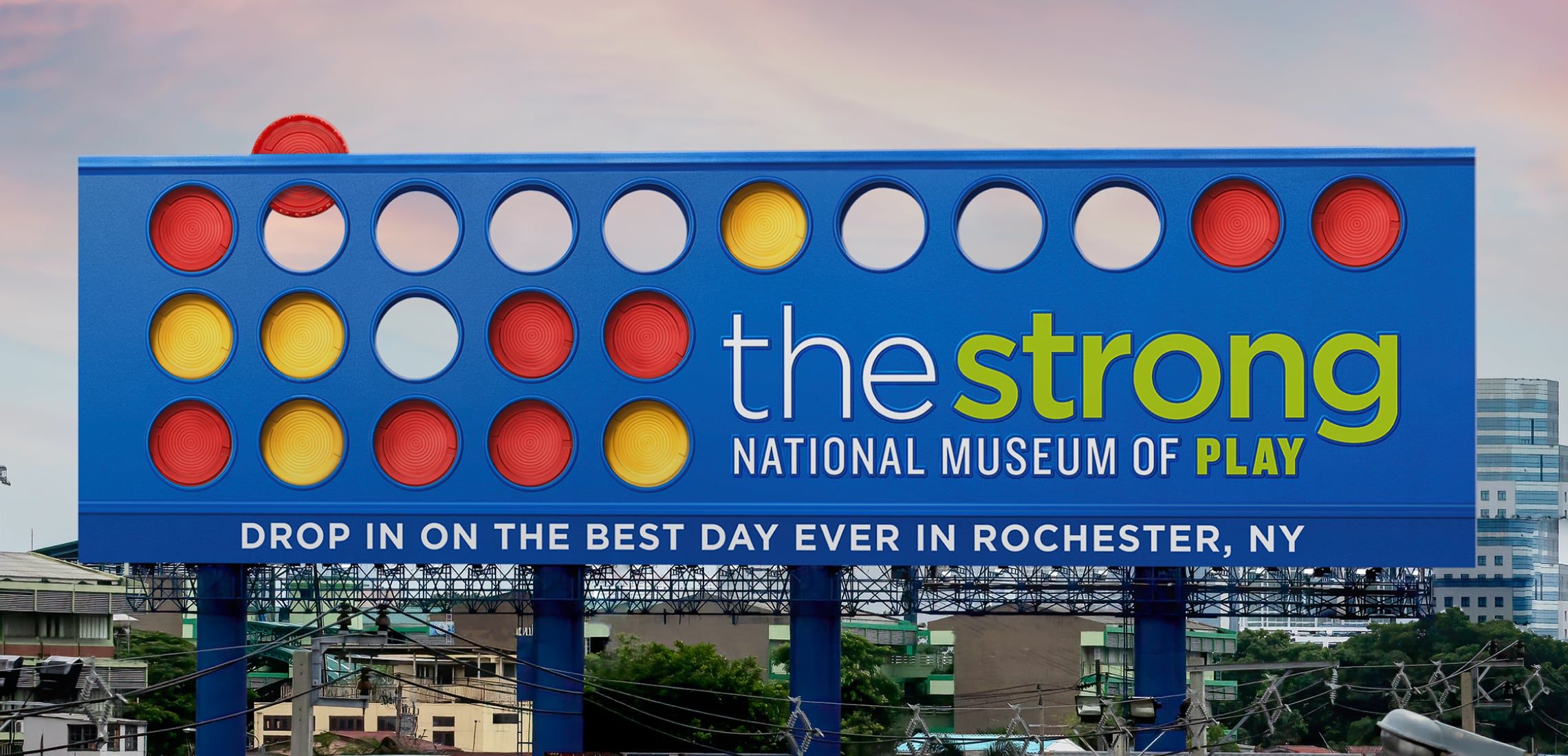 Photograph of a Strong National Museum of Play billboard that looks like the game Connect Four. It says, "Drop in on the best day ever in Rochester, NY."