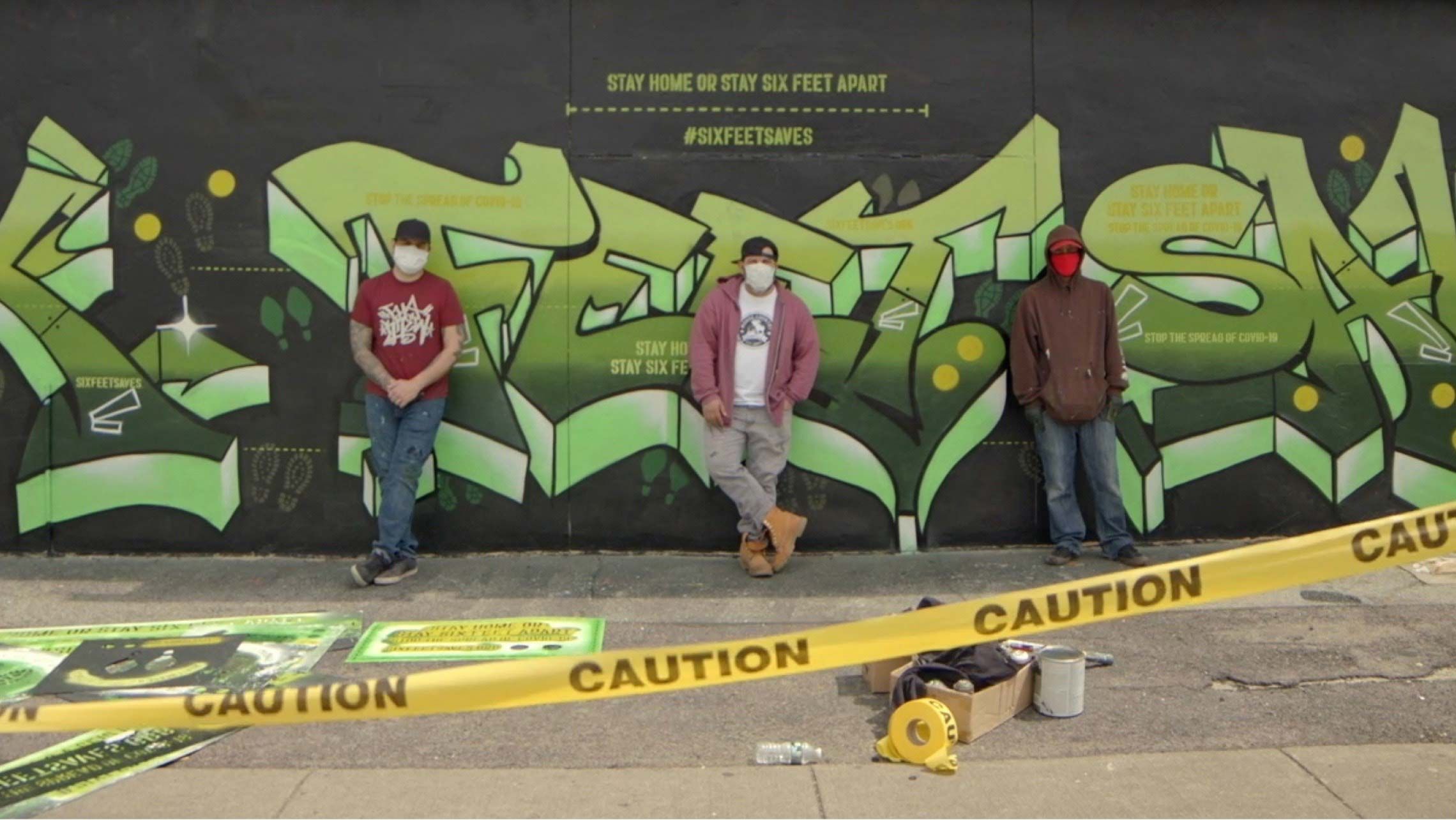 Image of the Six Feet Saves graffiti mural with the artists standing in front of it. Click to view the video about the Six Feet Saves mural.