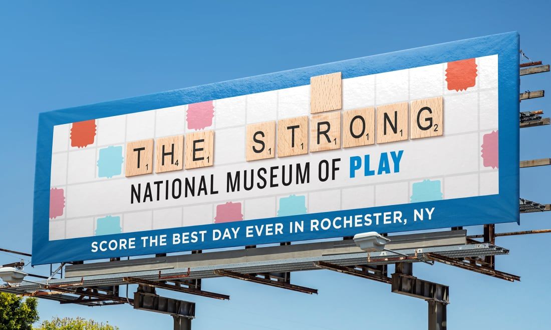 Photograph of a Strong National Museum of Play billboard that looks like a Scrabble board. It says, "Score the best day ever in Rochester, NY."