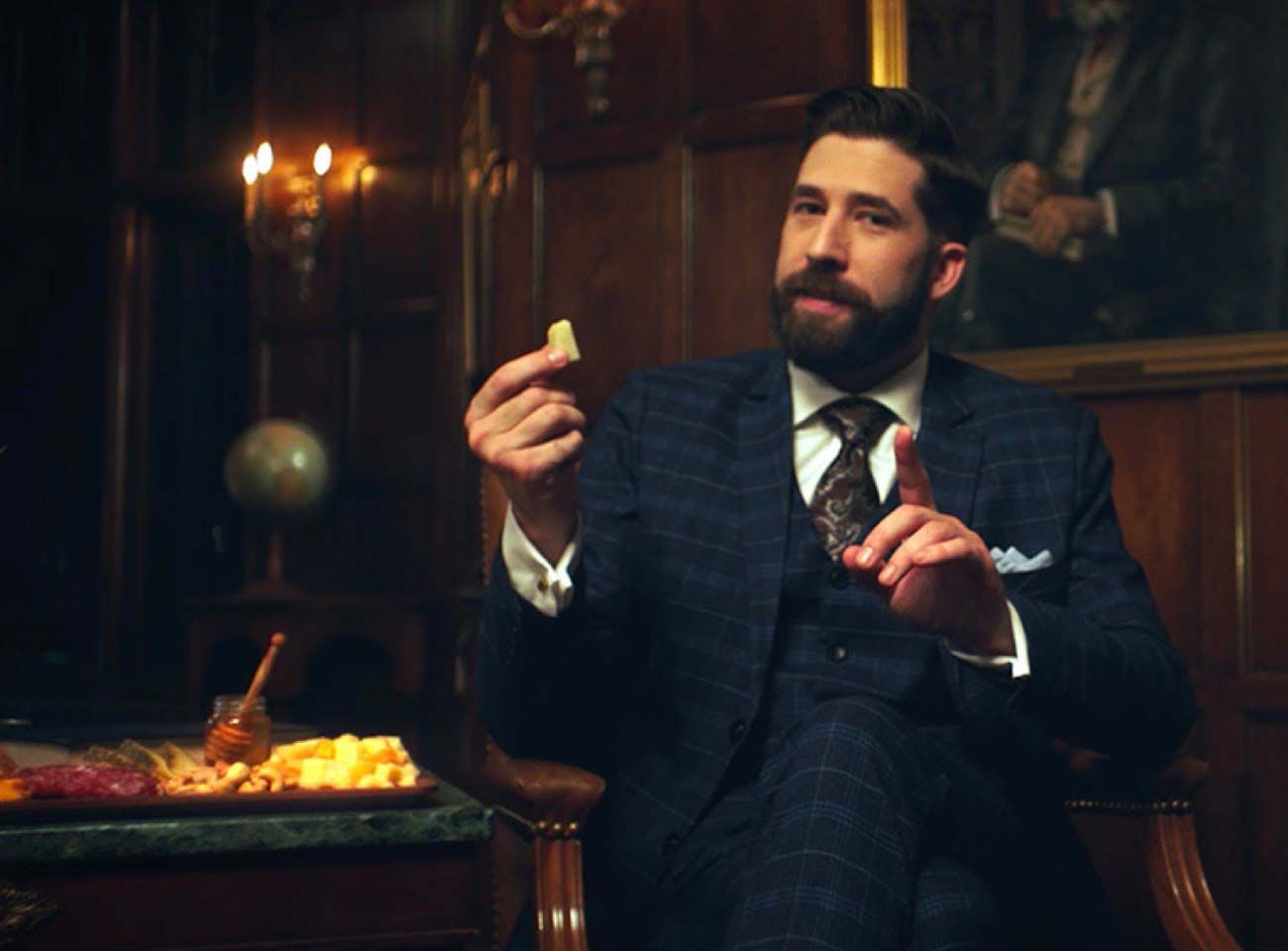 A well-dressed man sitting in a chair next to a platter of cheese. Click to view the video.