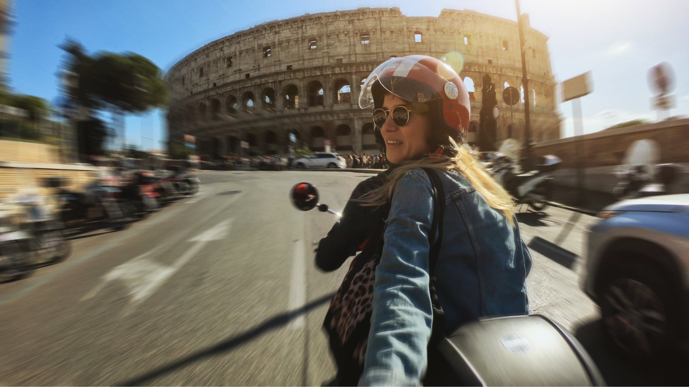 Image of a woman riding on the back of a moped, filming herself in front of the Colosseum