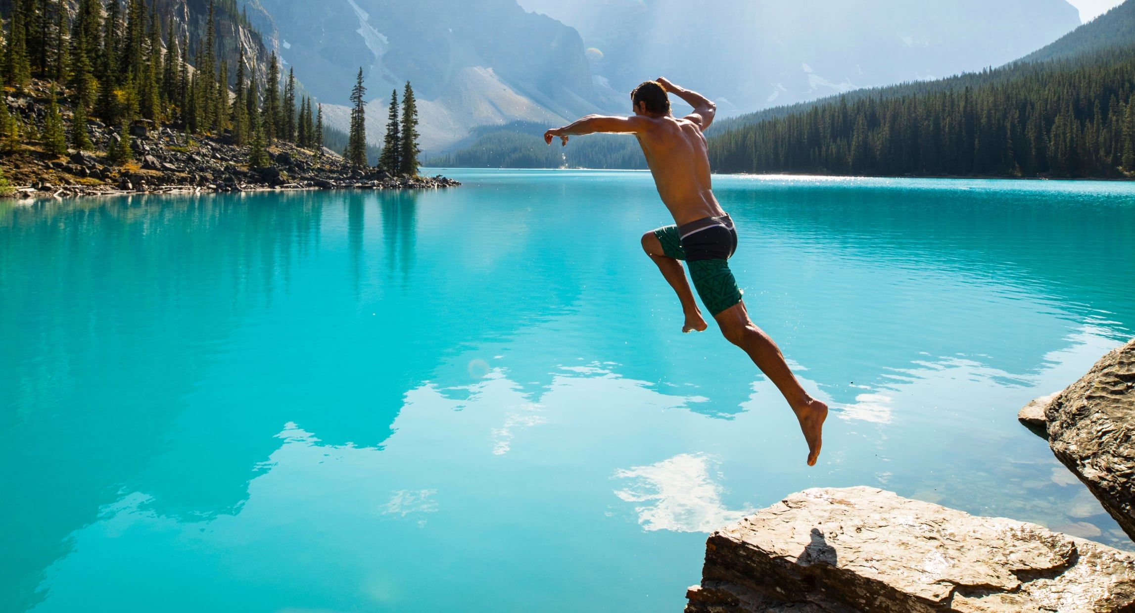 Photograph of a man jumping off of a rock into a gorgeous blue lake.