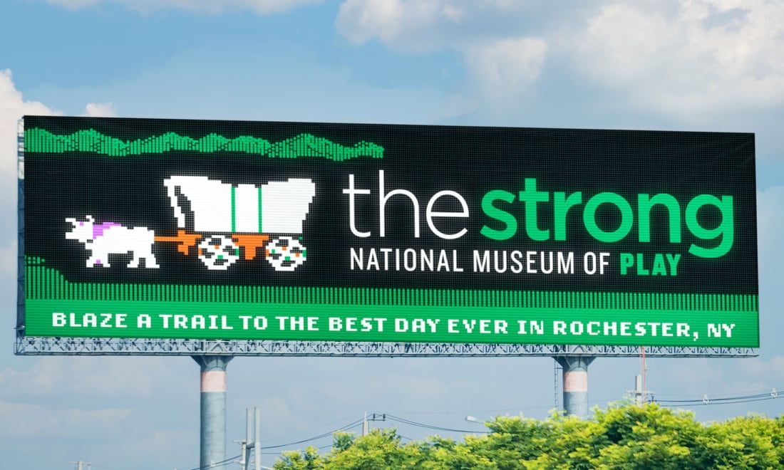 Photograph of a Strong National Museum of Play billboard that looks like the video game Oregon Trail. It says, "Blaze a trail to the best day ever in Rochester, NY."