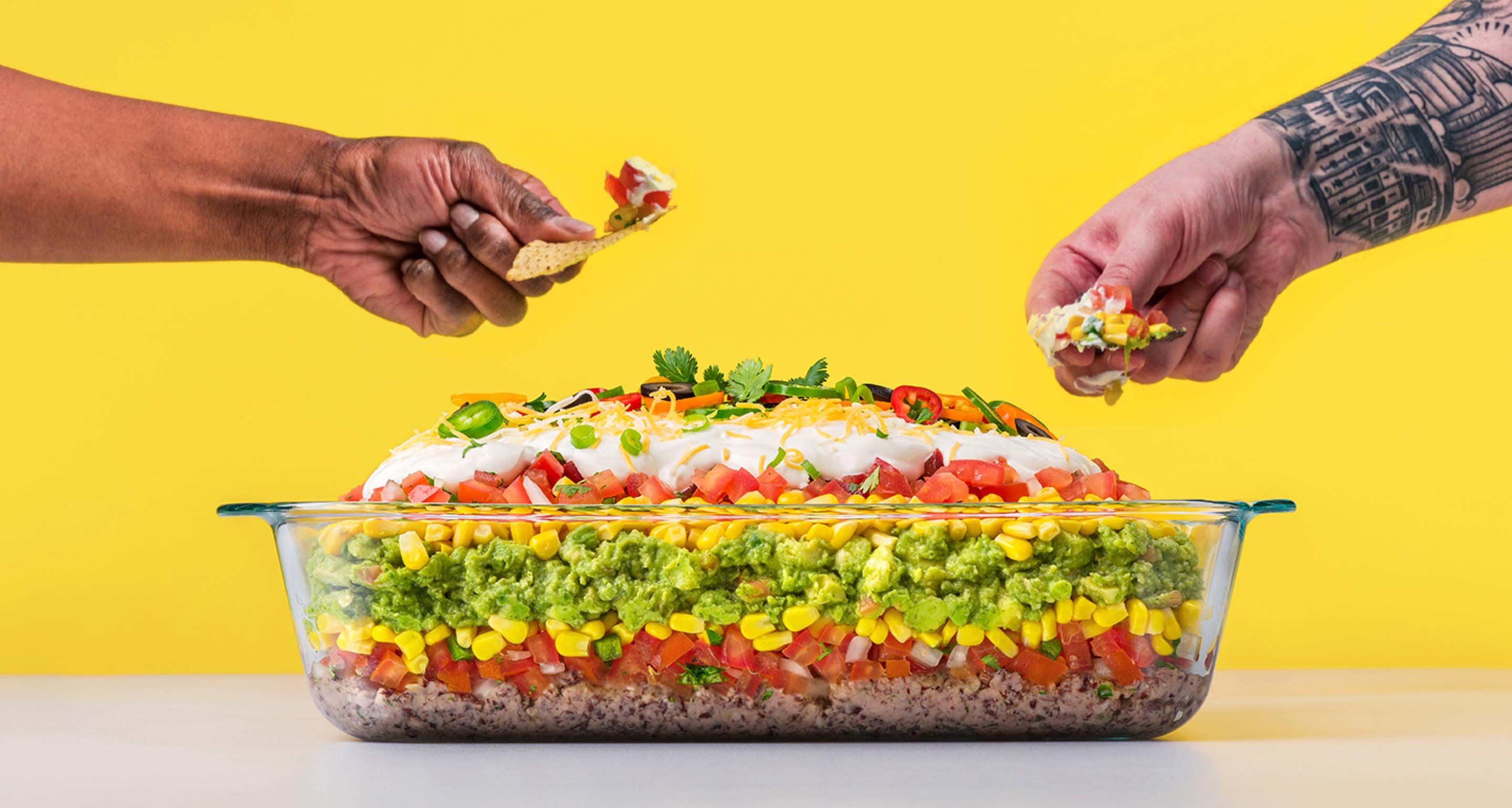 Photograph of two people dipping nachos in a Pyrex container of dip.