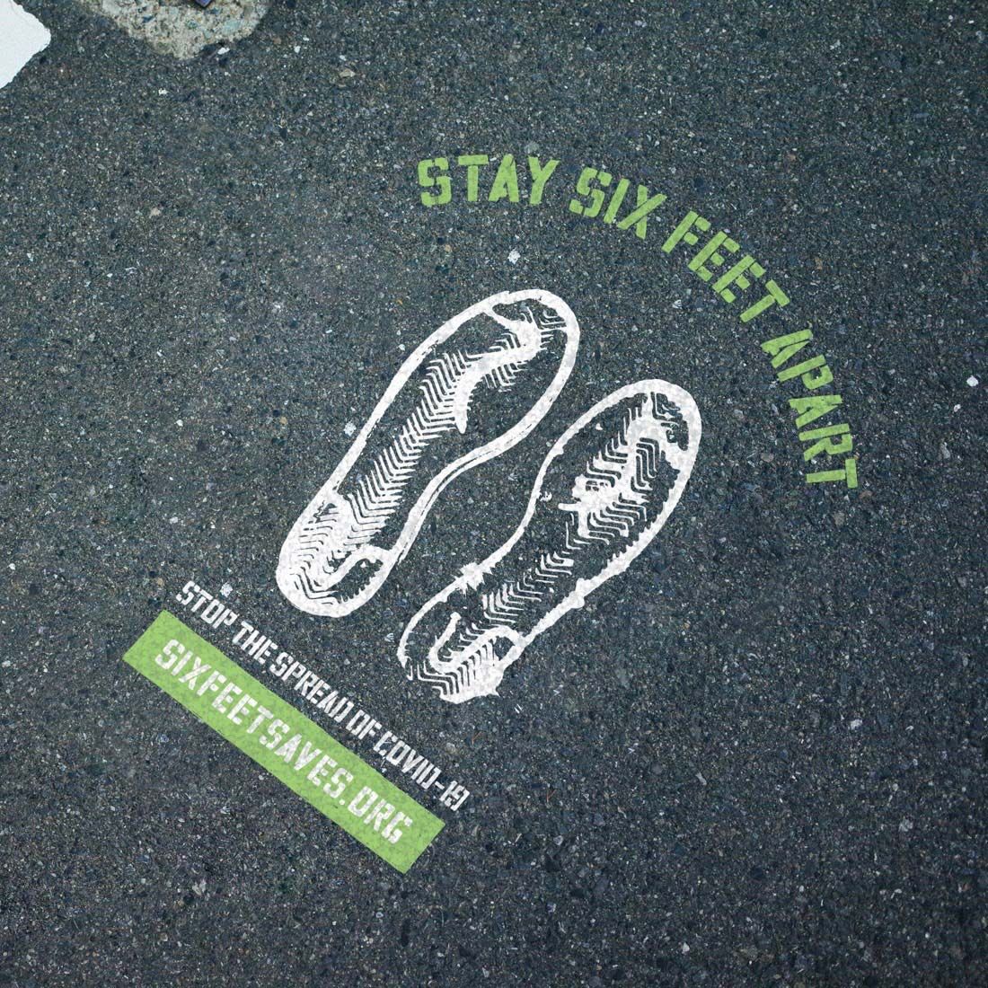 Photograph of a Six Feet Saves marker spray-painted onto the concrete, showing two footprints and saying, "Stay six feet apart."