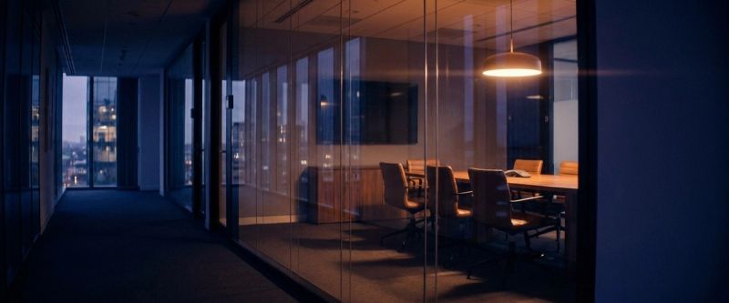 A lit glass-walled meeting room seen from a dark corridor