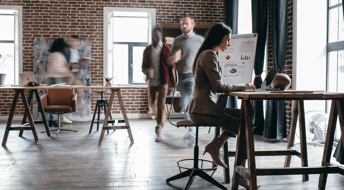 A woman works on a laptop at a desk in a modern office with a brick wall, while several people walk and talk in the background.