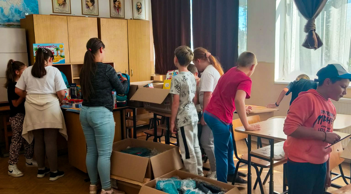 Children in a classroom gather around tables and open cardboard boxes filled with school supplies. The room is bright with natural light, creating an atmosphere of excitement and curiosity.
