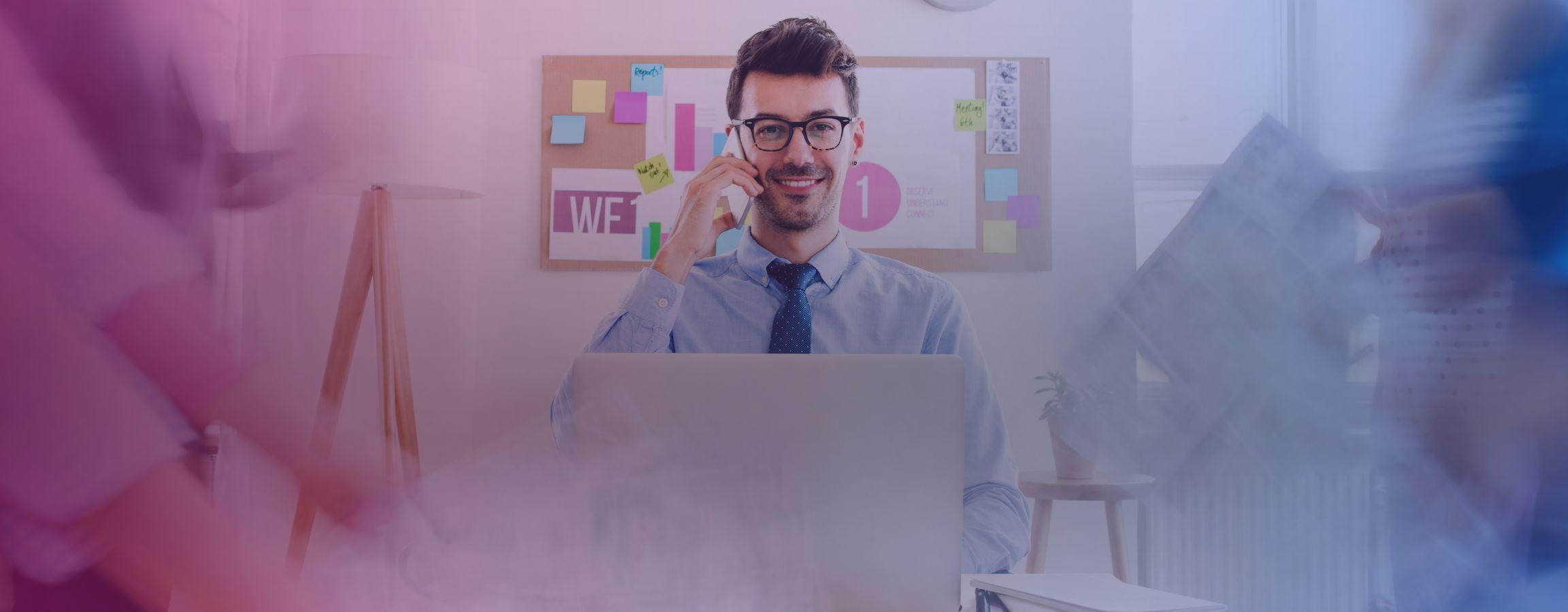 A business man on the phone whilst smiling at the camera and working on his laptop