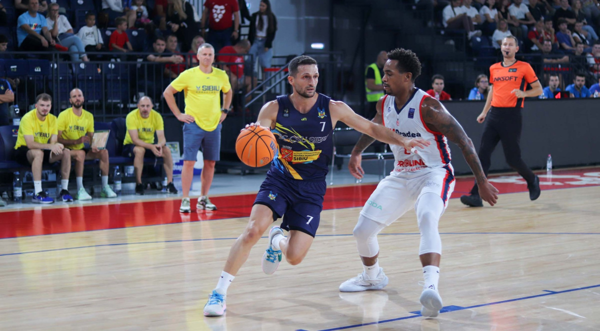 Two basketball players in action on the court, one dribbling the ball while being closely guarded, with spectators and team staff in the background.