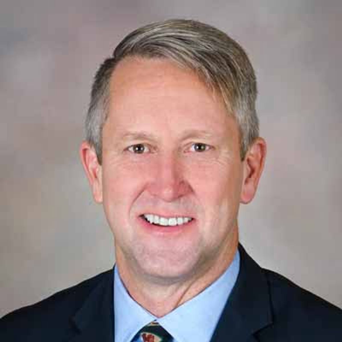 Professional headshot of middle-aged man with gray hair wearing dark suit and blue shirt with red tie against neutral background.
