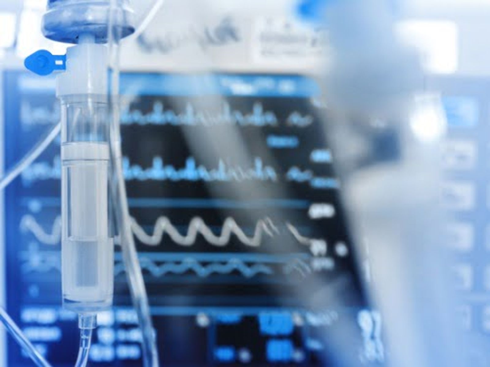 Laboratory test tube with liquid in foreground, medical monitoring equipment with waveform displays blurred in background.