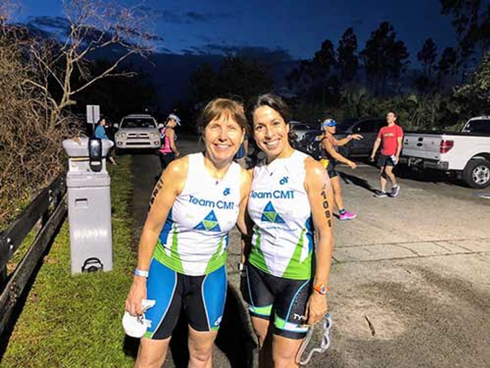 Two women in Team CMT athletic wear pose together at twilight event with parked vehicles and spectators visible.