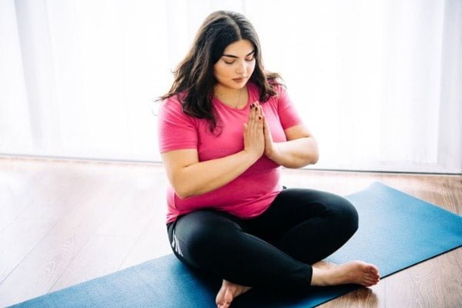 Woman in pink shirt and black leggings practicing yoga meditation on blue mat indoors.