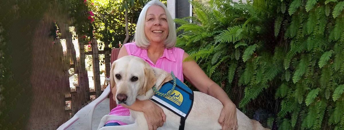 Woman in pink shirt with white service dog wearing blue vest in garden setting.