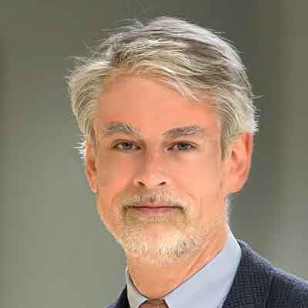 Retrato profesional de un hombre de mediana edad con cabello gris y vello facial, con camisa azul y corbata.
