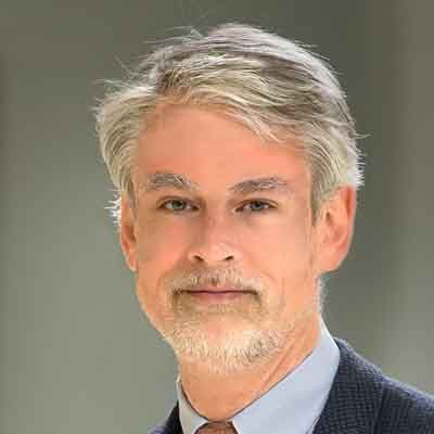Professional headshot of a middle-aged man with gray hair and facial hair wearing a blue shirt and tie.