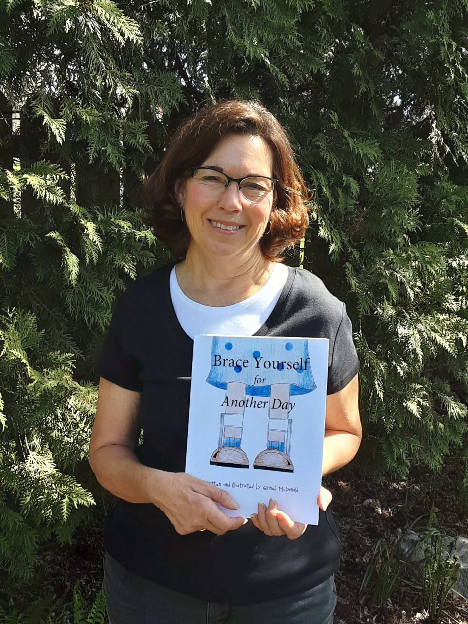 Mujer que sostiene el libro infantil "Brace Yourself for Another Day" sobre un fondo de árboles de hoja perenne