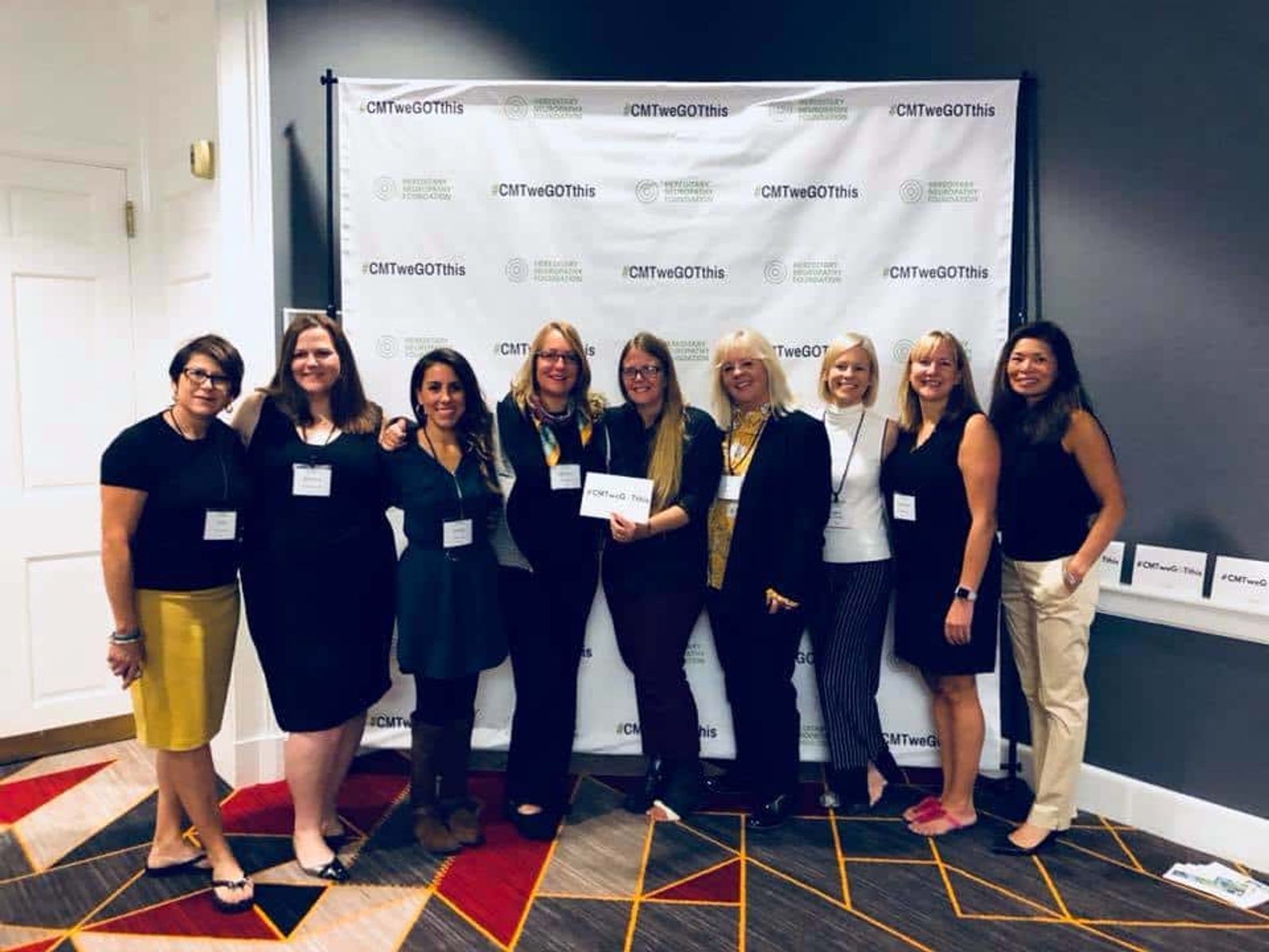 Ten women standing together in front of branded backdrop at professional event with name badges.