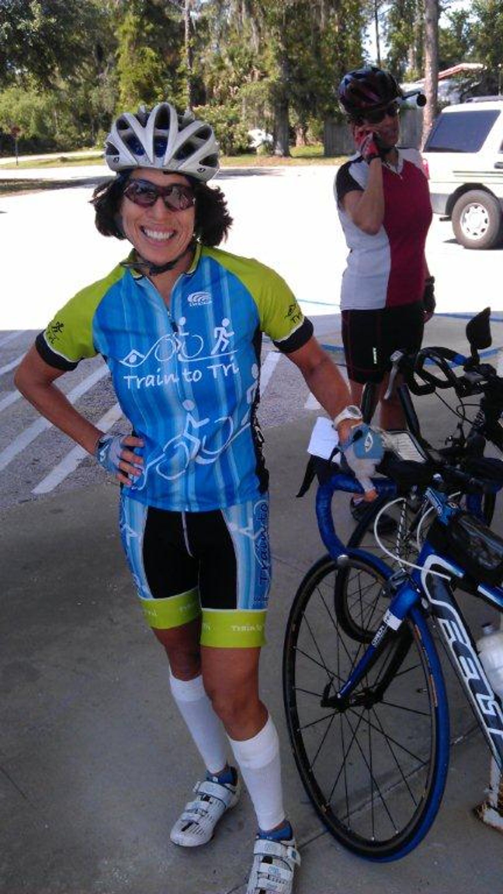 Cyclist in blue and green "Train to Tri" jersey with helmet standing next to road bike outdoors.