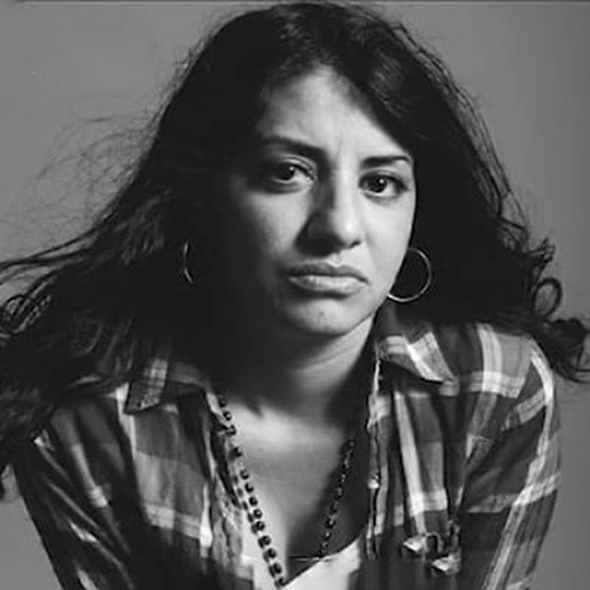Retrato en blanco y negro de una mujer con cabello oscuro largo, que lleva una camisa a cuadros y aretes de aro.