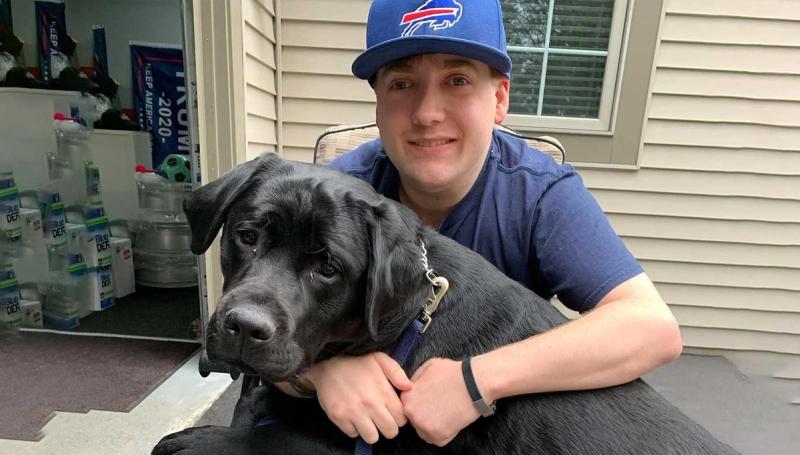 Person wearing Bills baseball cap posing with black dog indoors.