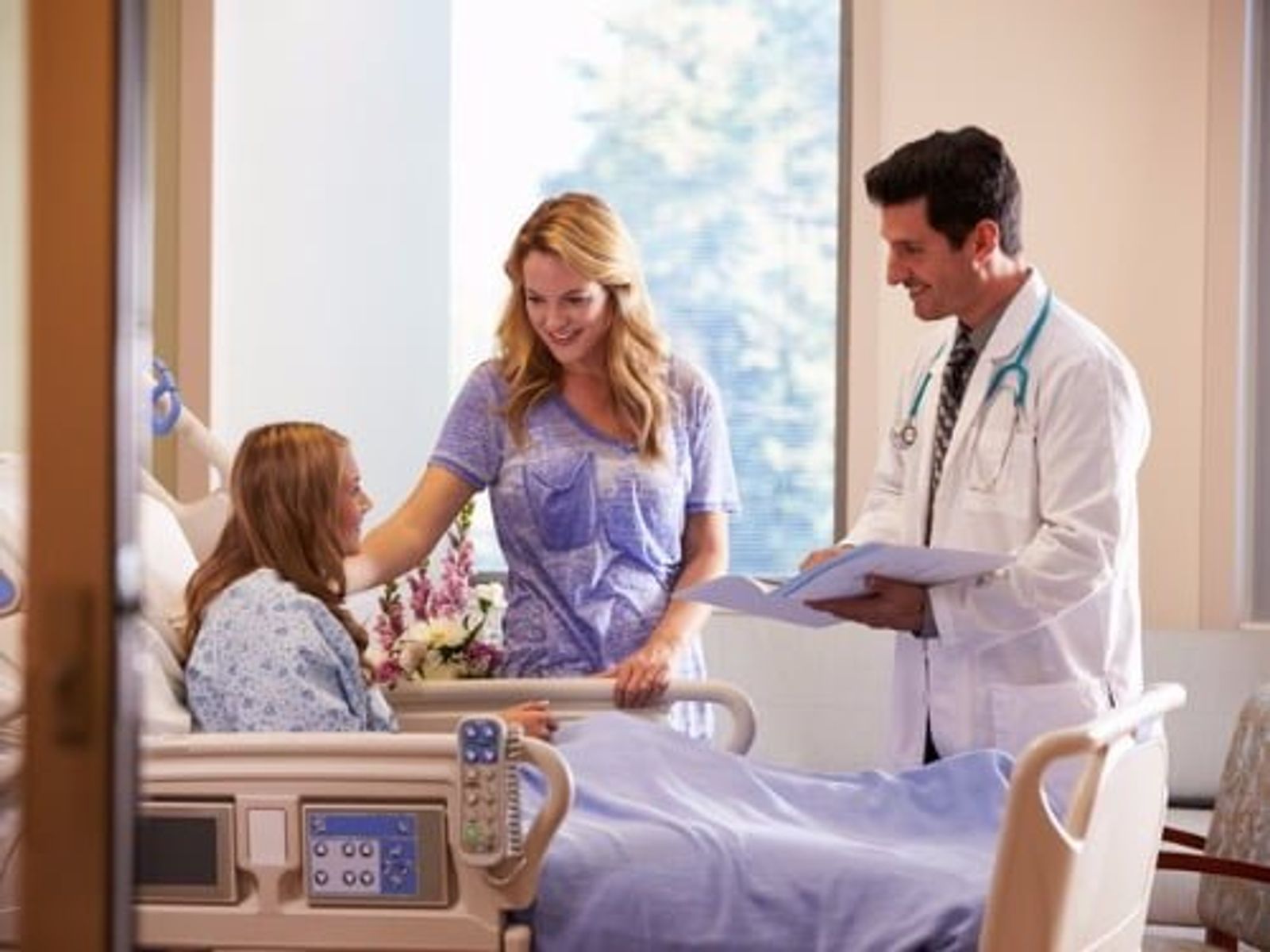 Doctor in white coat speaks with young patient and woman in hospital room.