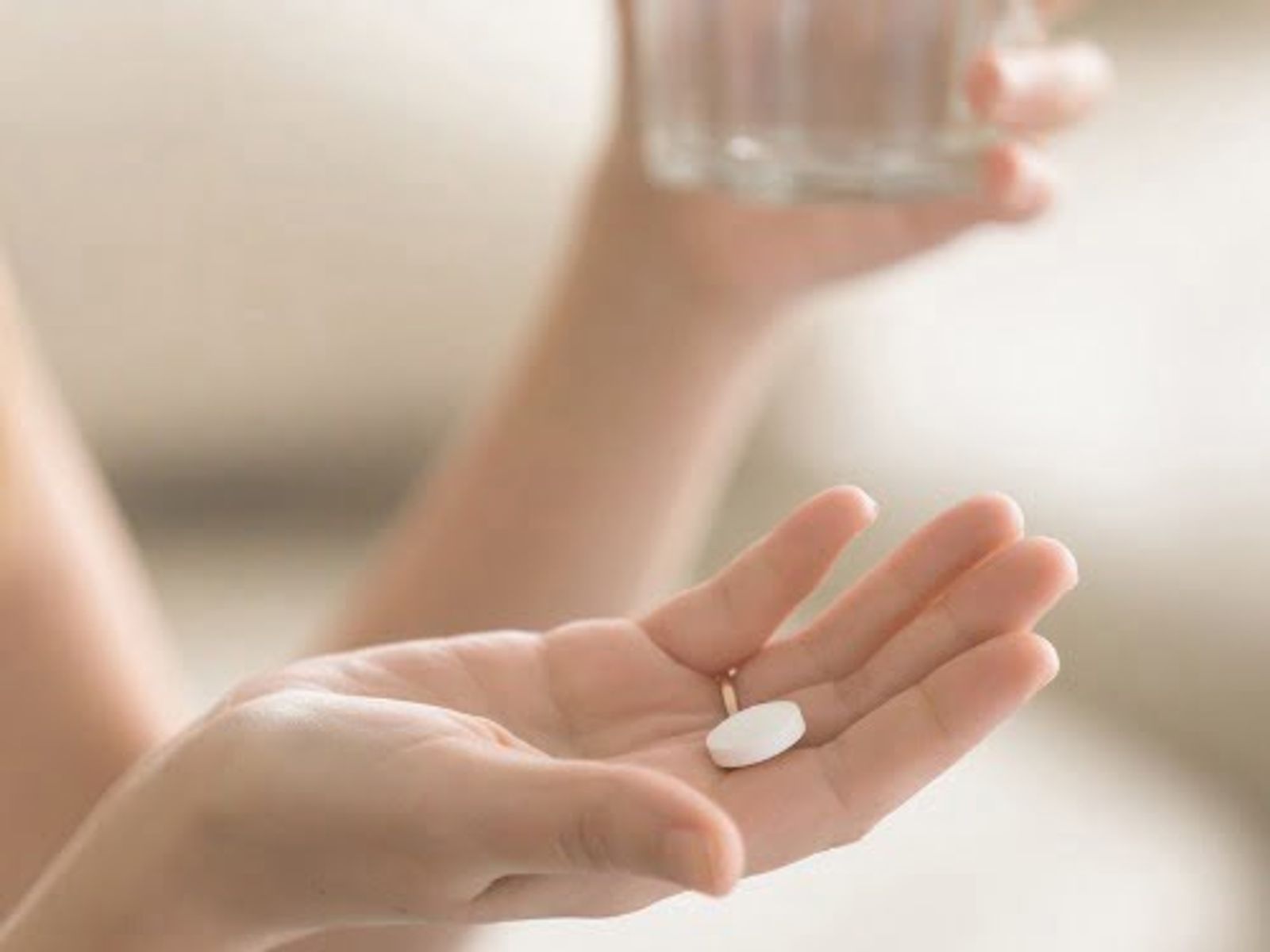 Persona sosteniendo una píldora blanca en la palma de la mano, con un vaso de agua al fondo.