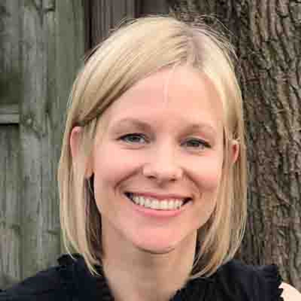 Blonde woman smiling at camera, wearing dark top, tree trunk background.