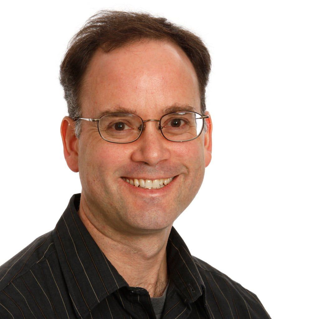 Professional headshot of a man wearing glasses and a dark pinstriped shirt, smiling at camera.