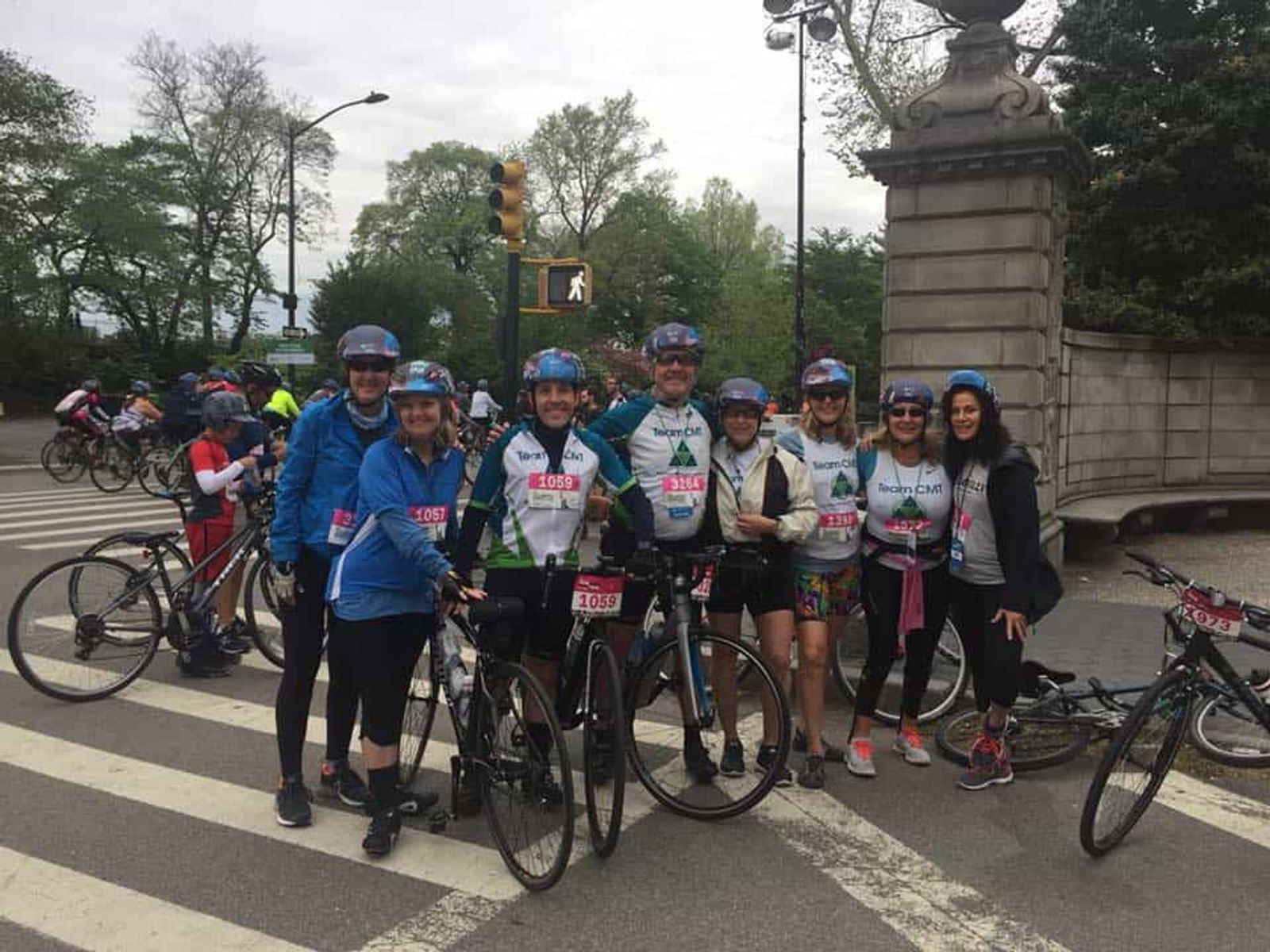 Group of cyclists wearing blue helmets and race bibs gathered on city street with monument.