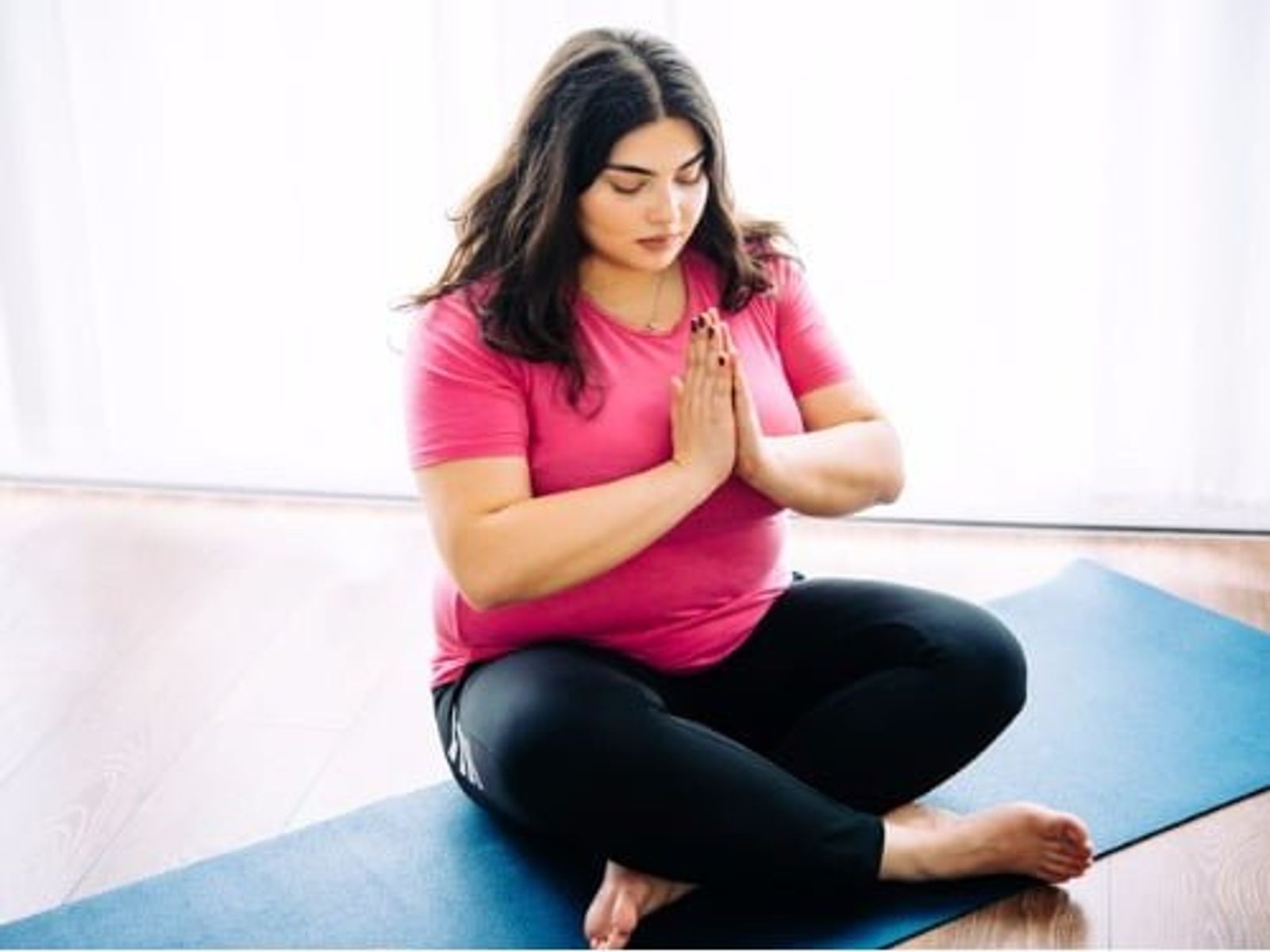 Woman in pink shirt and black leggings practicing yoga meditation on blue mat indoors.