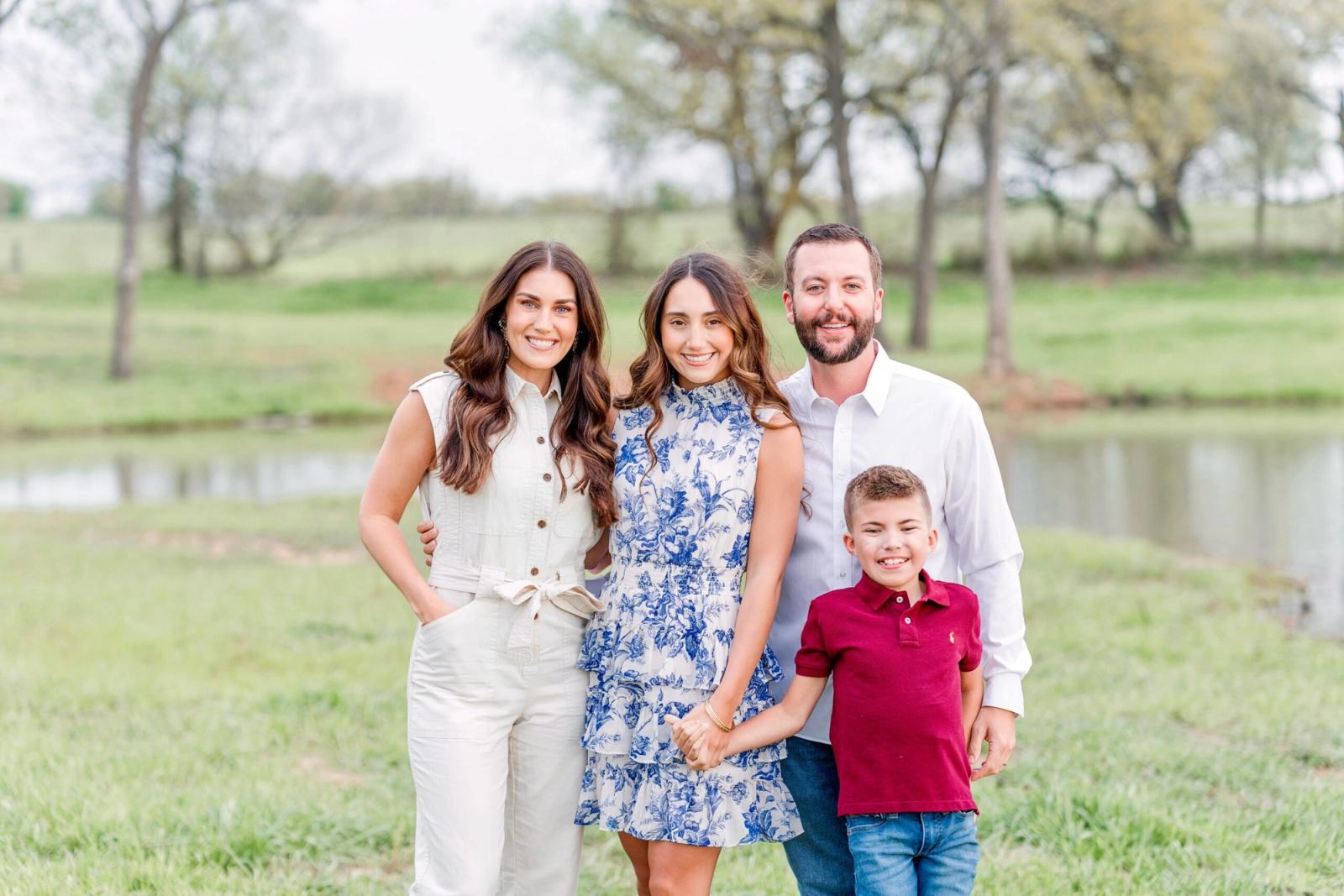 Family of four smiling outdoors in park setting with water and trees behind them.