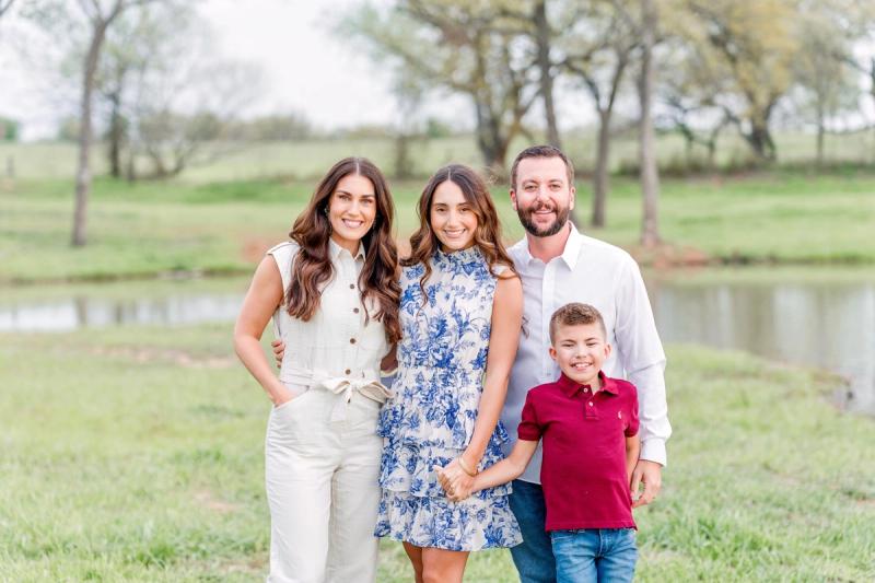 Family of four smiling outdoors in park setting with water and trees behind them.