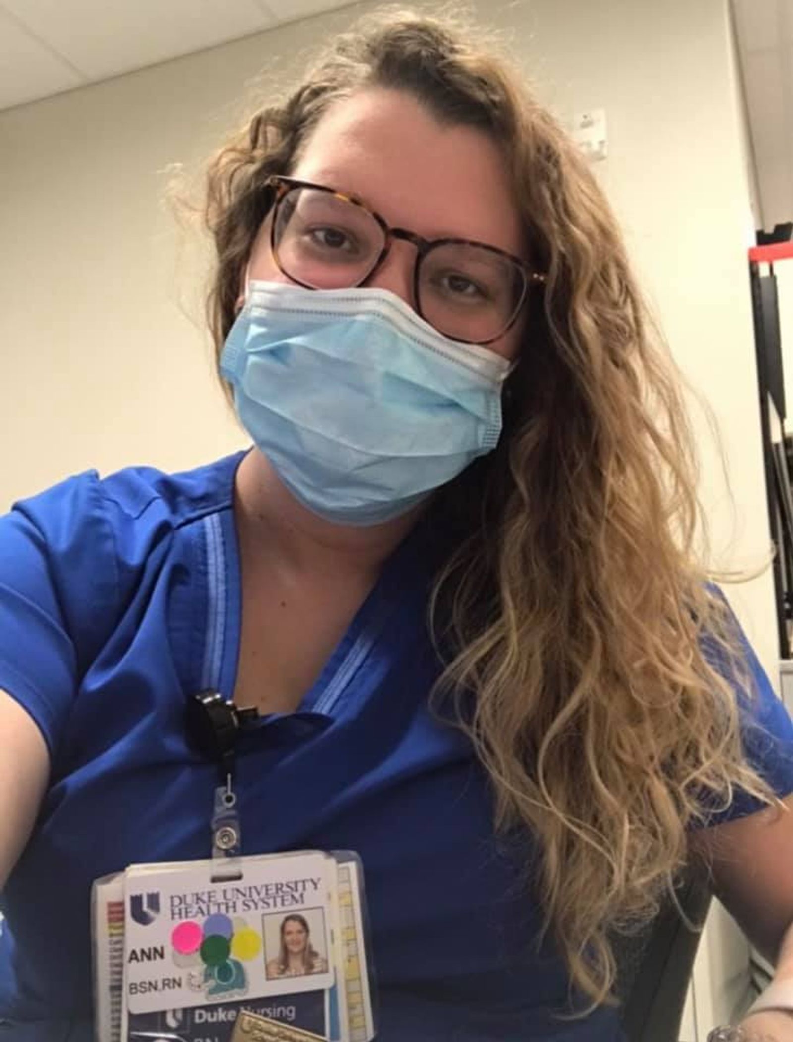 Healthcare worker wearing blue scrubs, mask, and Duke University hospital ID badge.