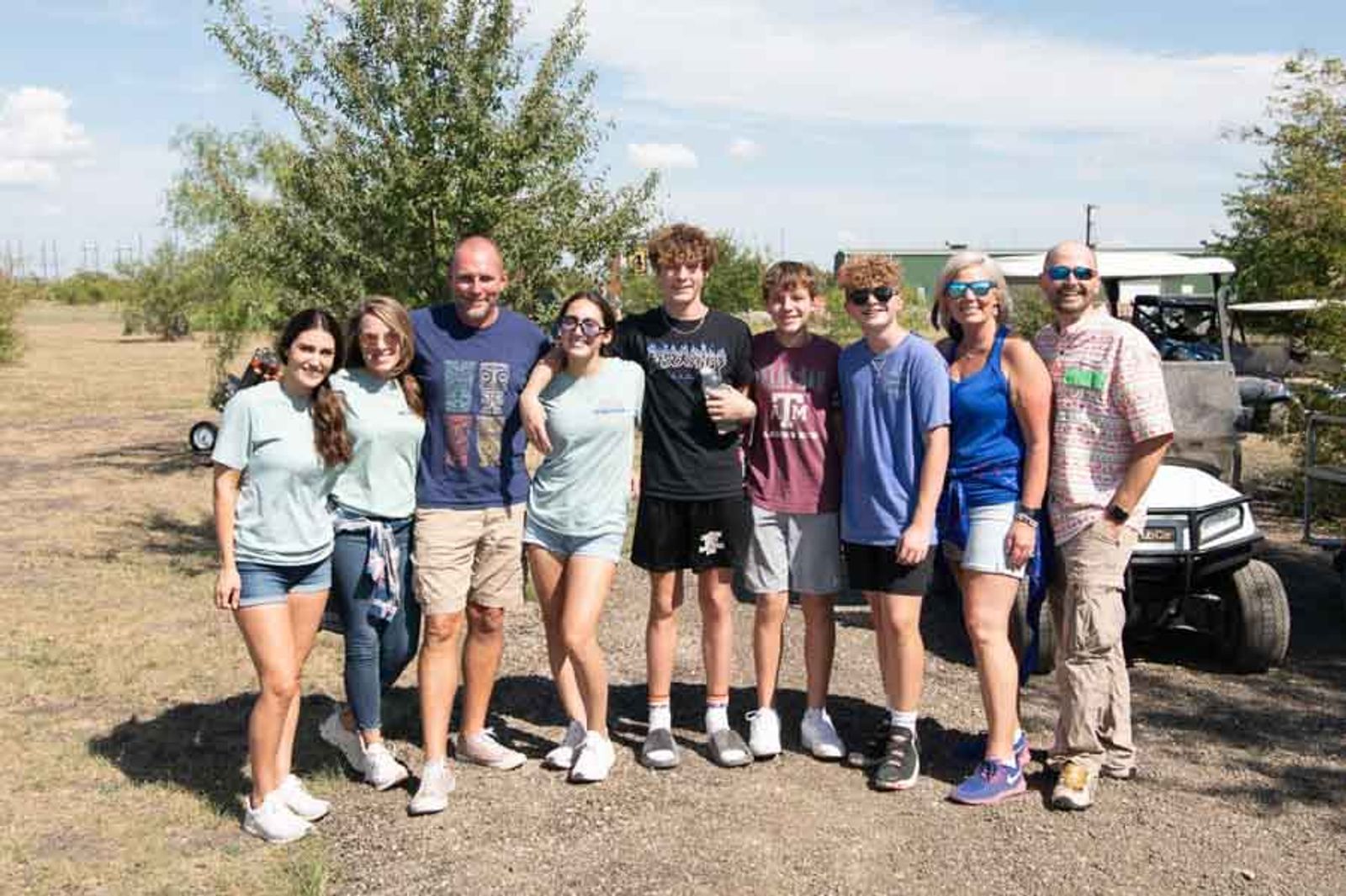 Group of twelve people standing together on rural property with golf carts and trees in background.