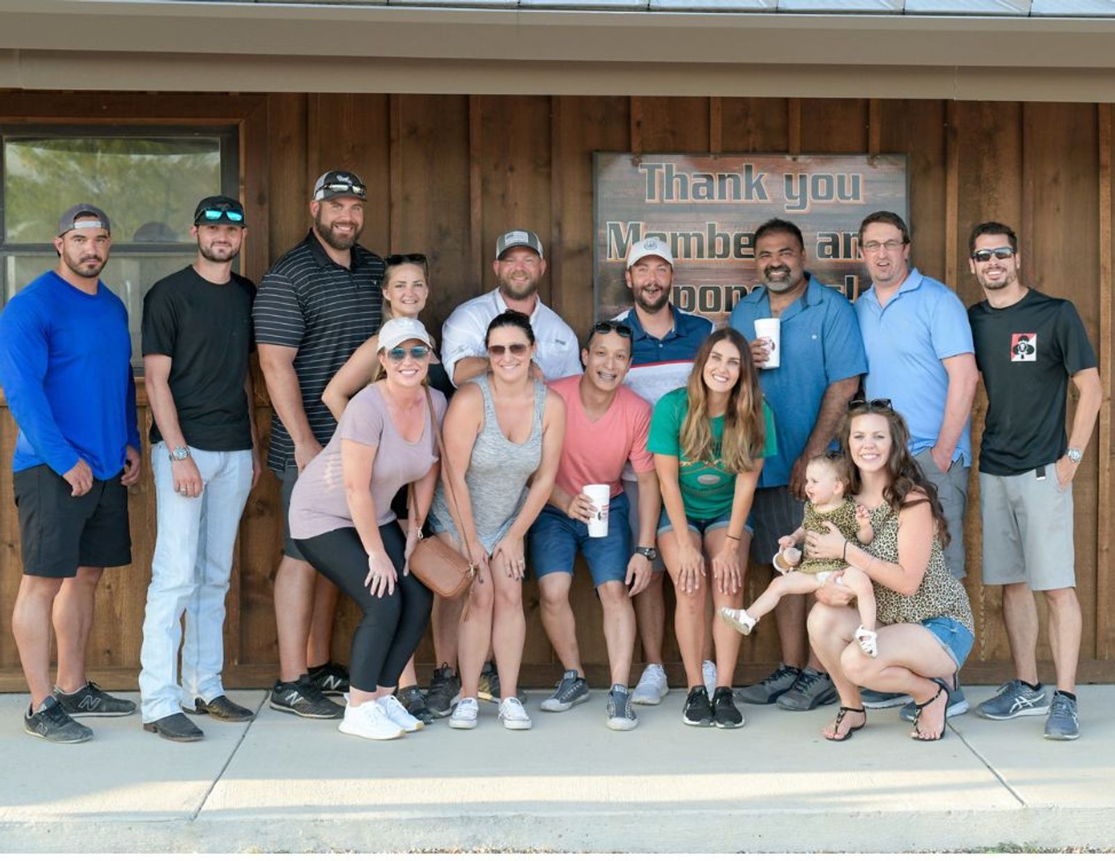 Group of approximately 16 people posing together outside wooden building with "Thank you Members for your support" banner.