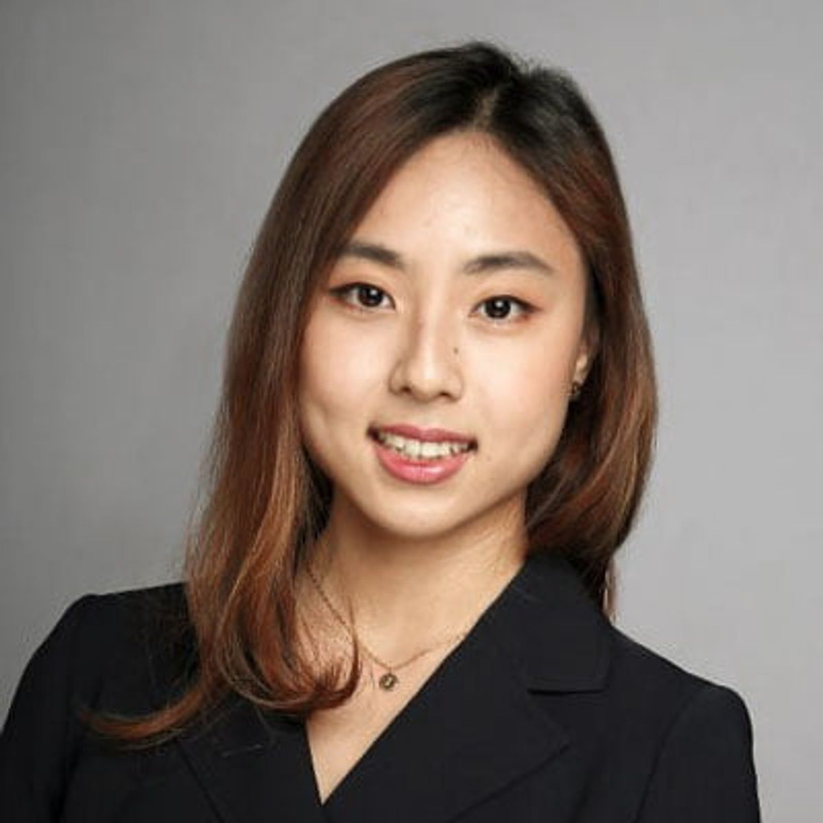 Professional headshot of a woman with long brown hair wearing a black blazer against a gray background.