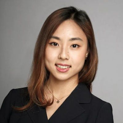 Retrato profesional de una mujer con cabello castaño largo que lleva un blazer negro sobre un fondo gris.