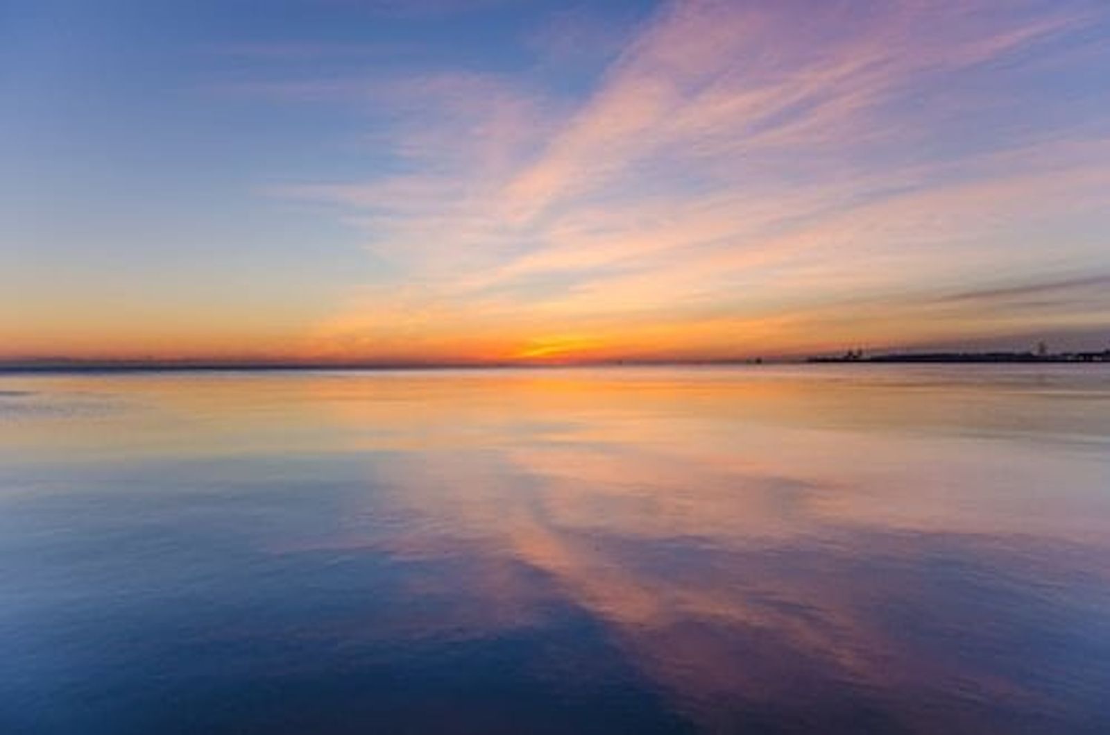Sunrise or sunset over calm water with colorful sky reflections and distant shoreline.