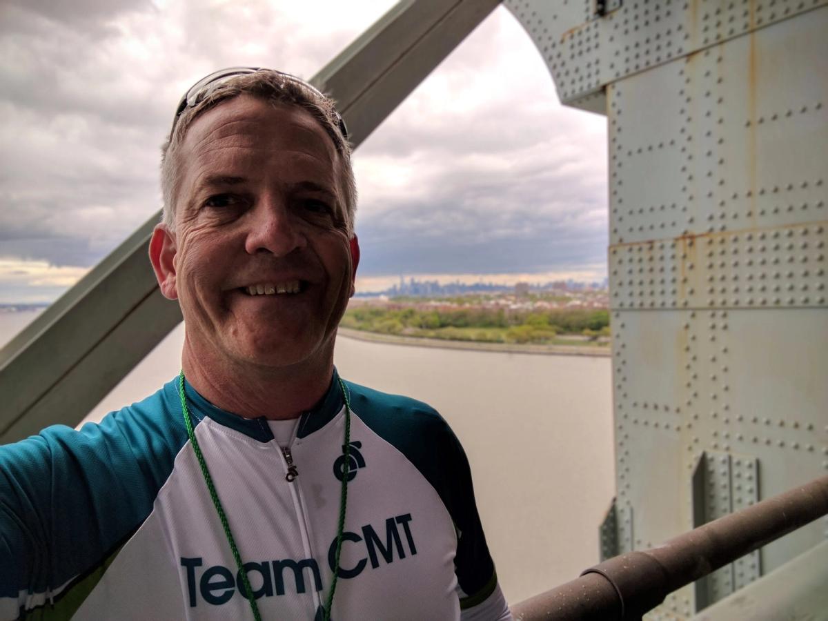 Man wearing Team CMT shirt smiling on bridge overlooking river and landscape