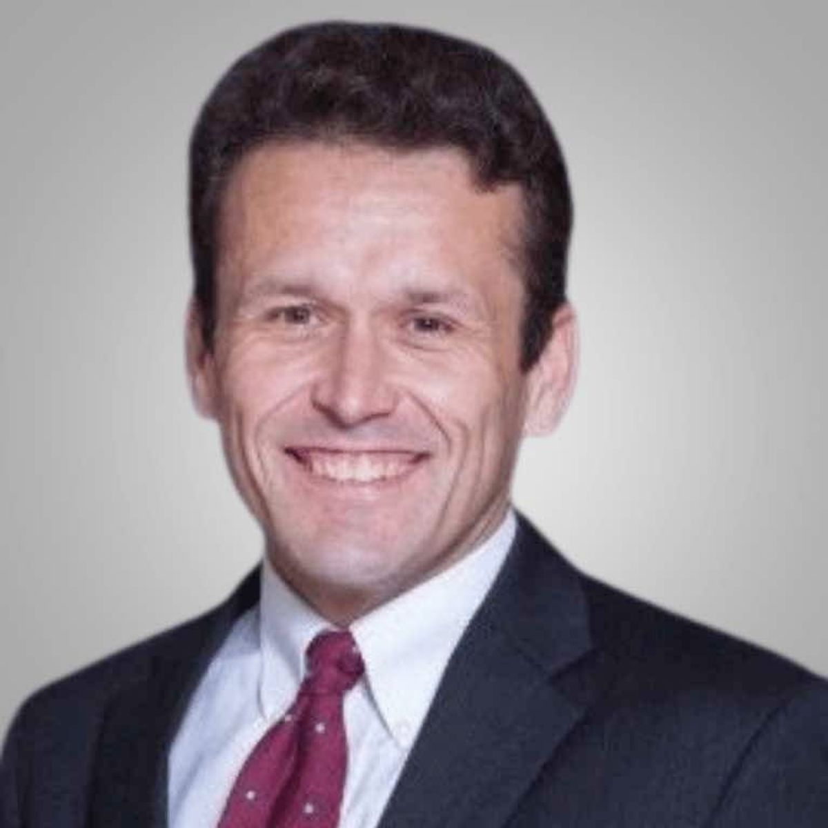 Professional headshot of a man in dark suit, light blue shirt, and maroon tie, smiling against a gray background.