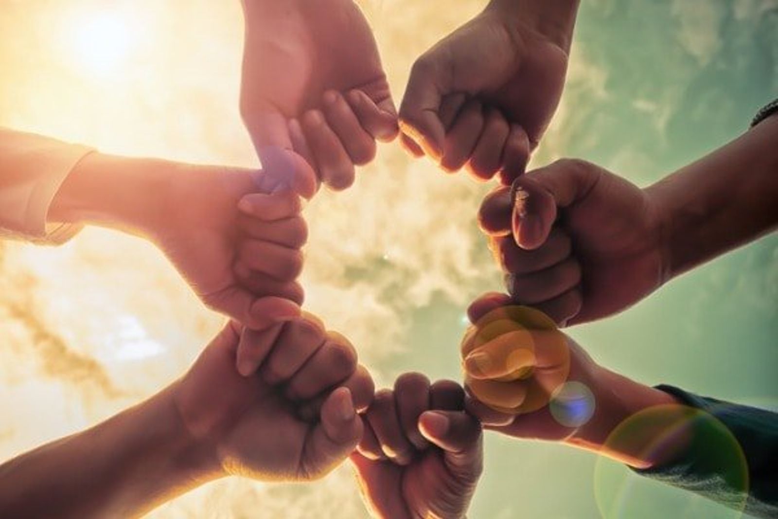 Multiple hands forming a circle together against bright sunlight and sky.