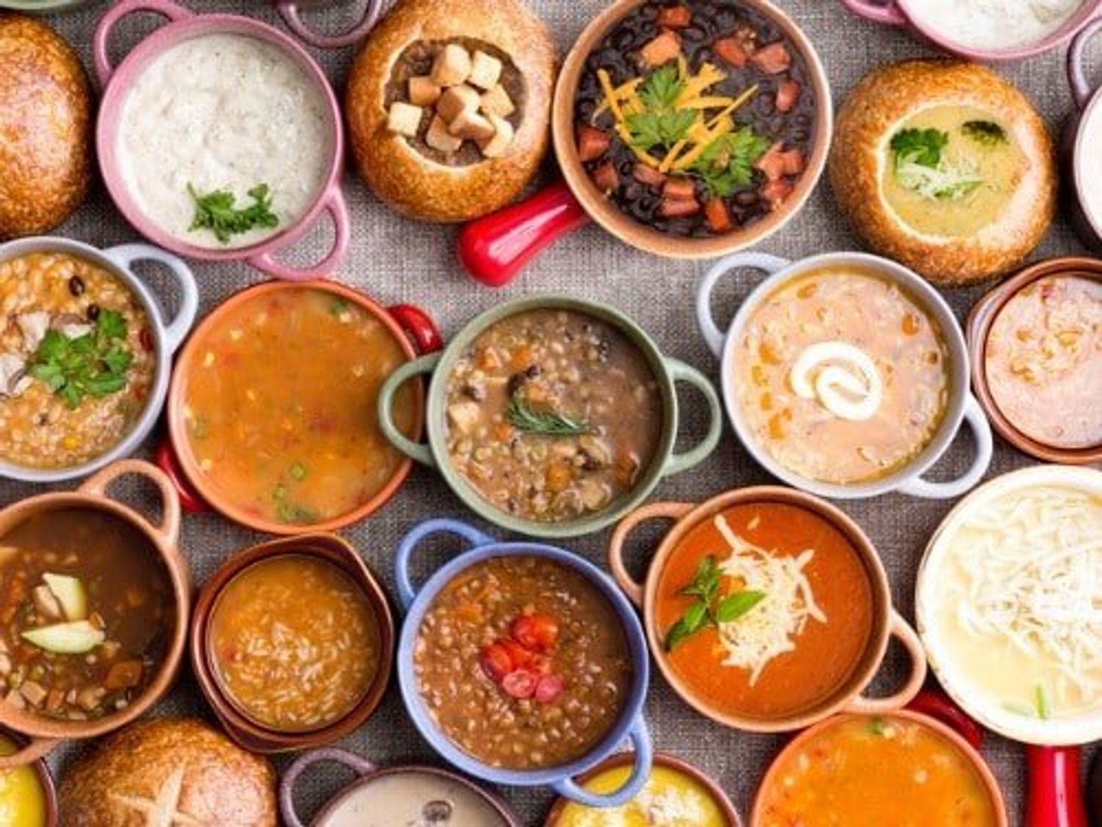 Overhead view of various soups and breads arranged on gray linen surface.