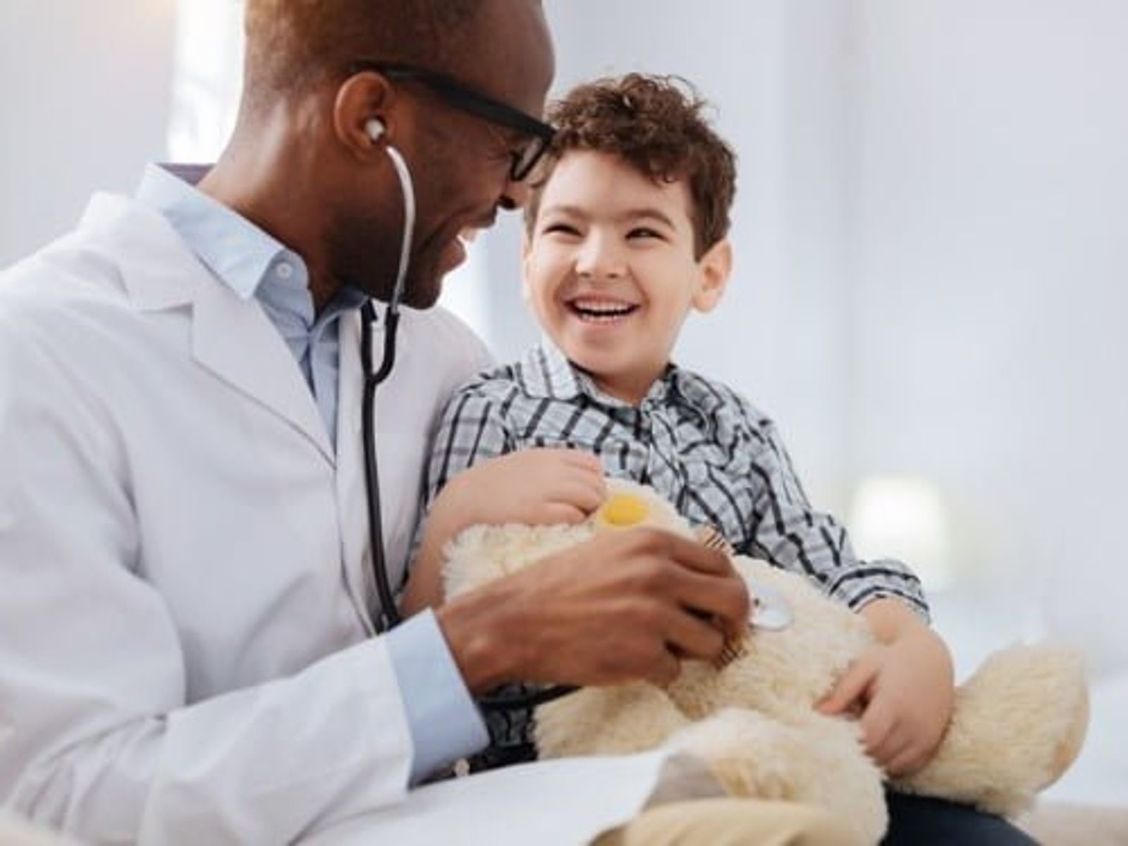 Médico con estetoscopio que examina a un niño pequeño que sostiene un osito de peluche durante un control médico.