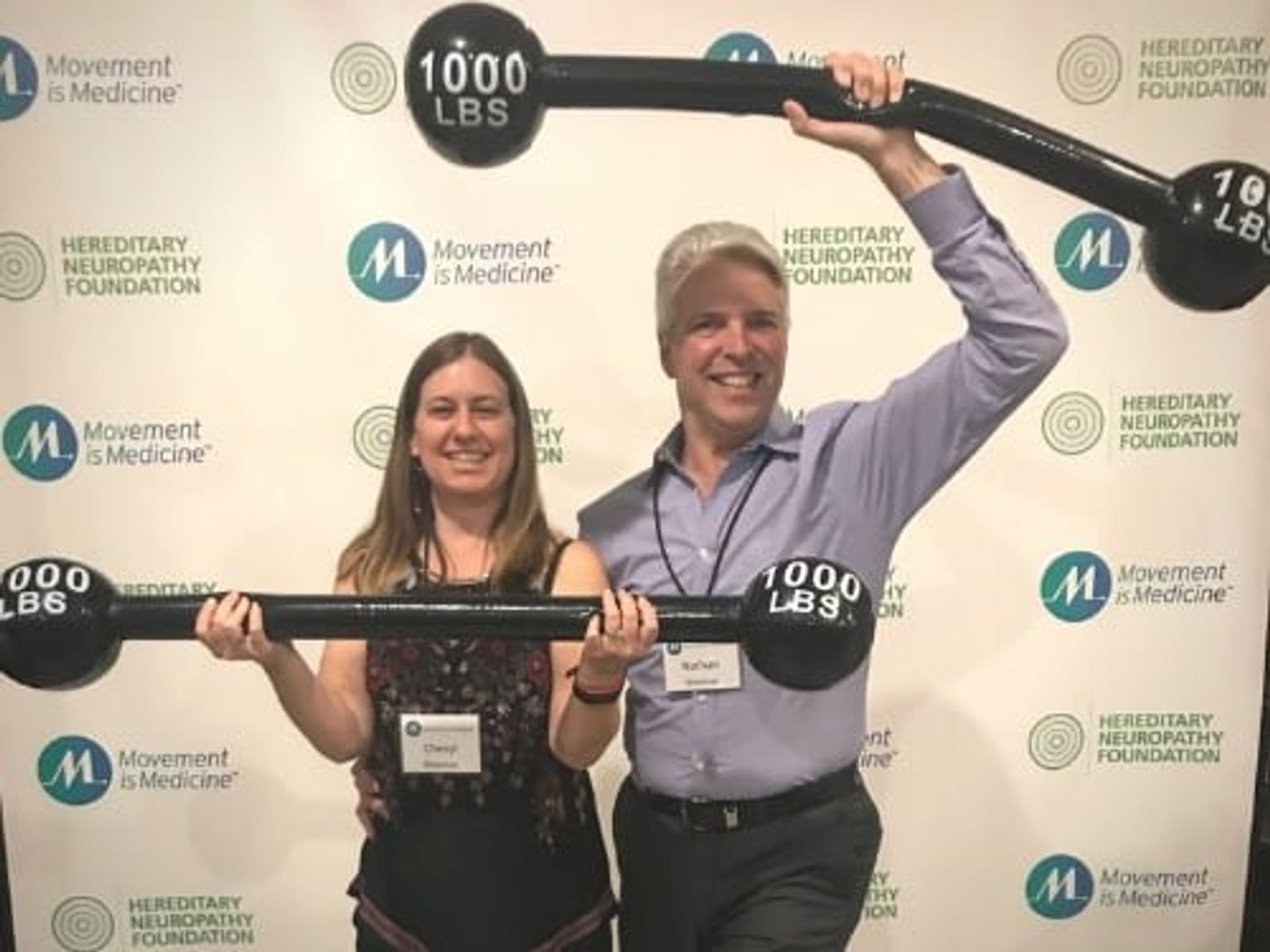 Two people holding weighted barbells at Movement is Medicine event backdrop.
