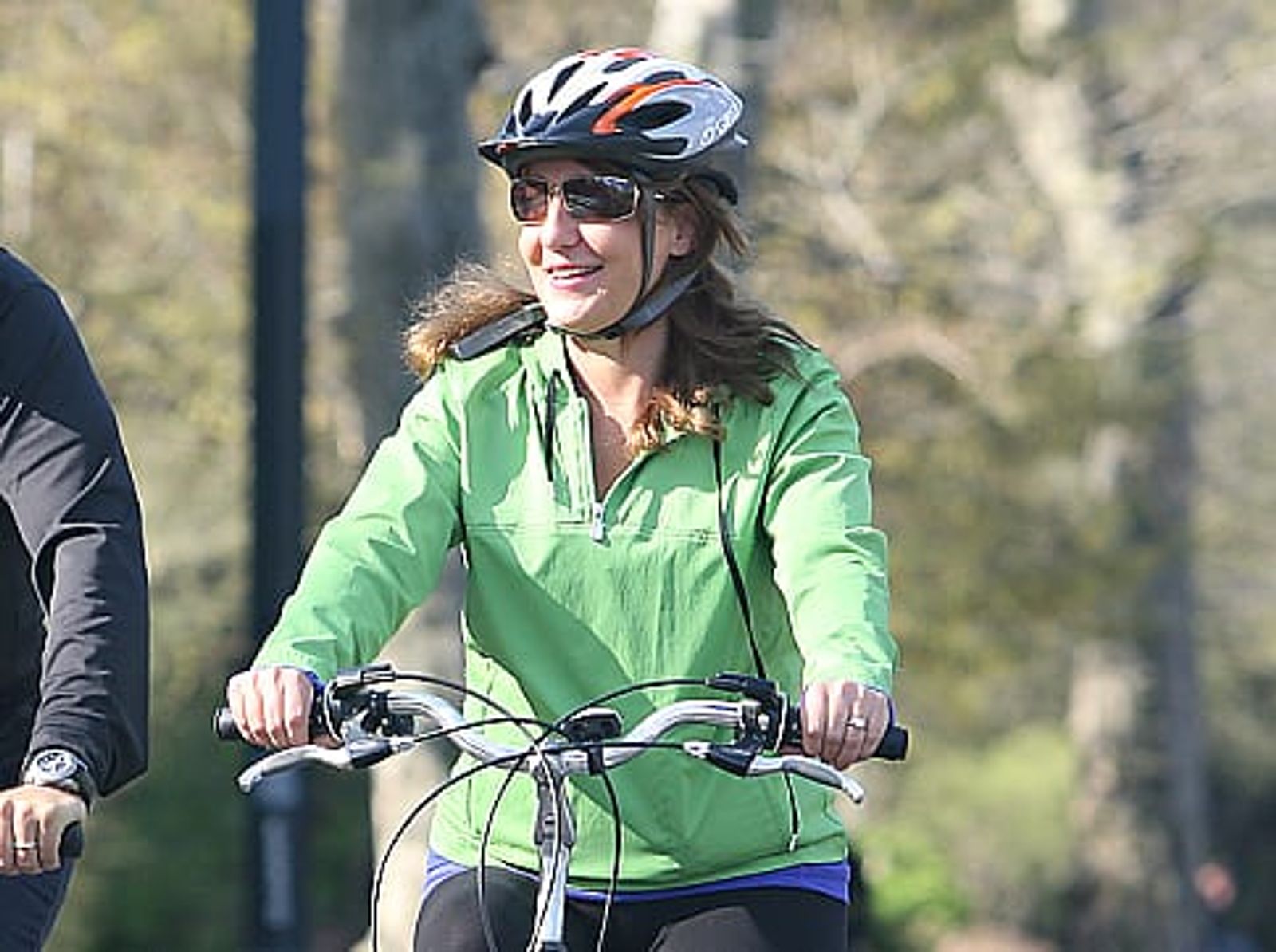 Woman in green jacket riding bicycle wearing helmet and sunglasses on tree-lined path.