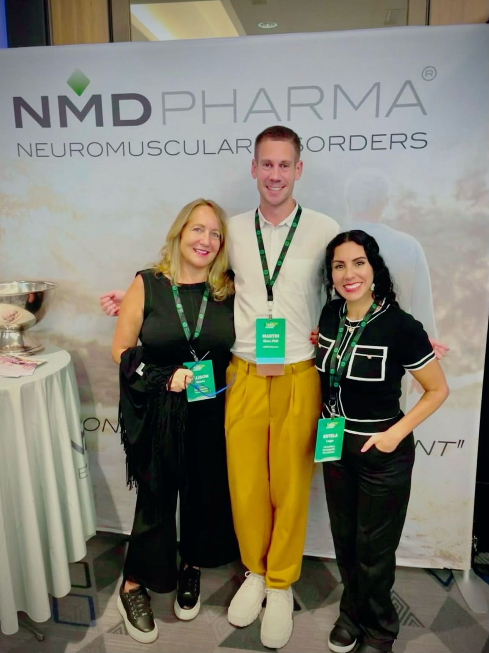 Tres personas con credenciales verdes de conferencia están frente a una pancarta de NMD Pharma Neuromuscular Disorders.