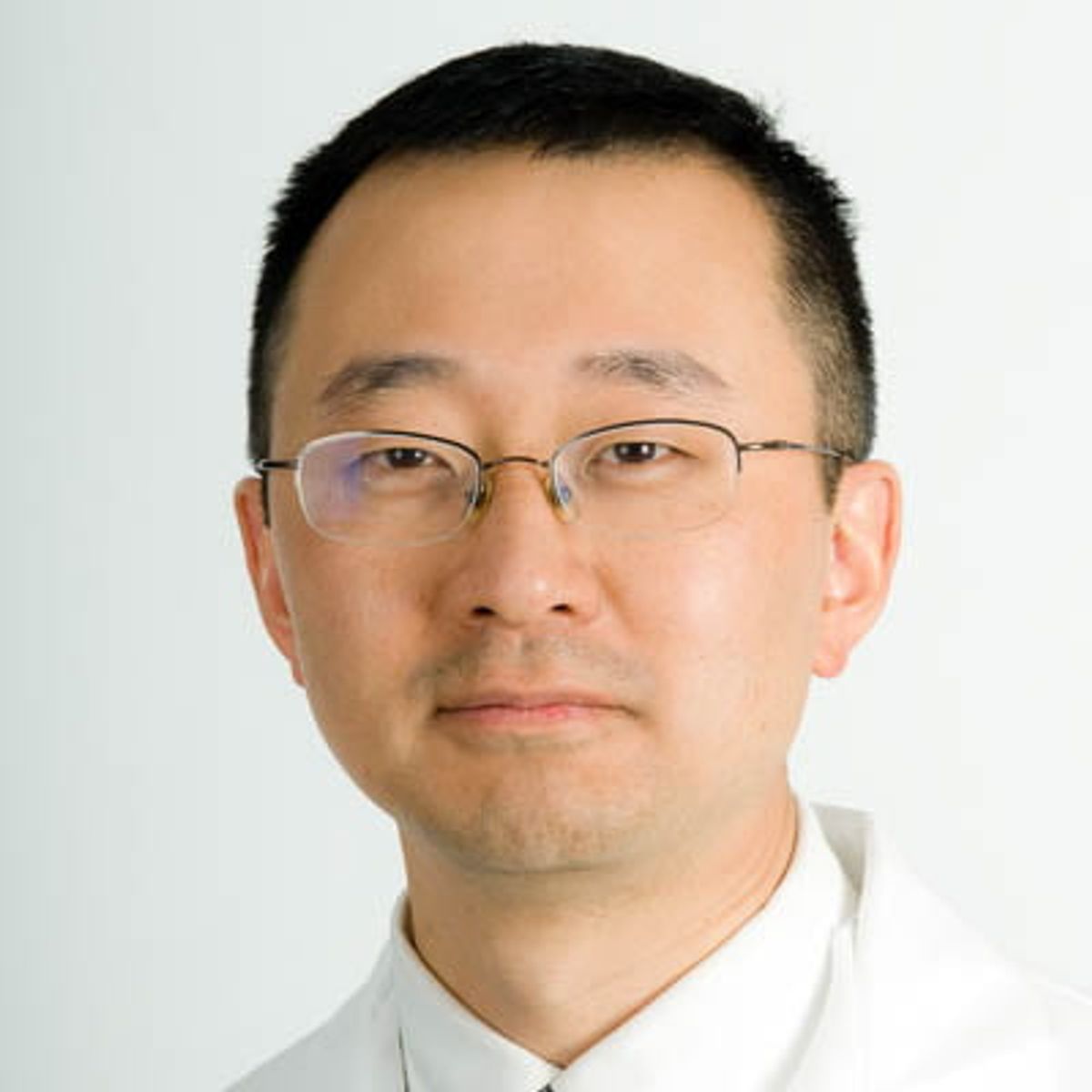 Retrato profesional de un hombre asiático con gafas y camisa blanca sobre un fondo claro.